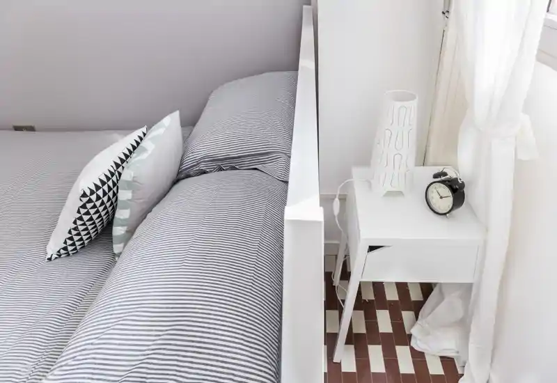 Habitación luminosa y minimalista con una cama doble bien hecha con ropa de cama a rayas, cojines decorativos y una mesita blanca con lámpara y despertador.