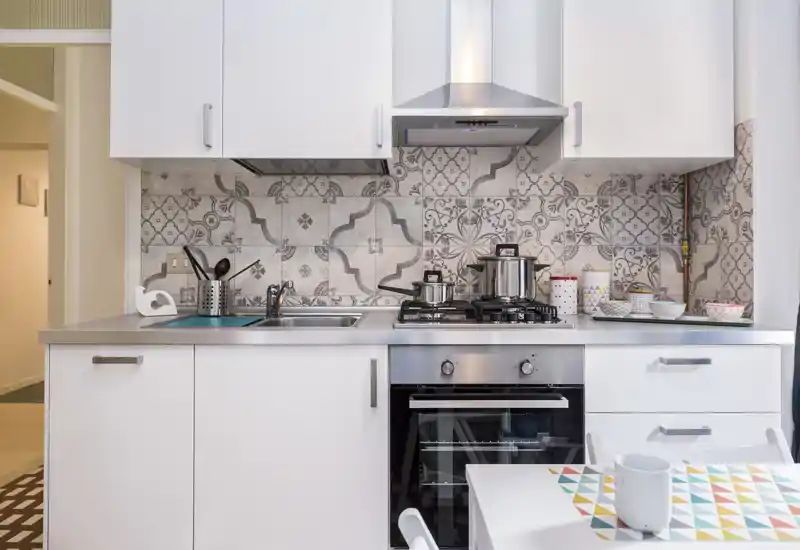 Modern compact kitchen with a stainless-steel oven and gas hob, decorative patterned backsplash, overhead extractor and neatly arranged cookware—bright and well-styled for short-term rentals.