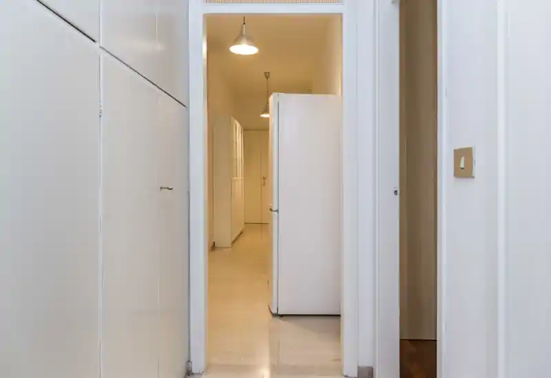 Narrow entrance corridor with built-in cupboards and a view into the hallway; neutral lighting and clean white finishes.