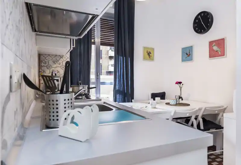 Modern compact kitchen with a countertop sink and stove, a small adjacent dining table set for two, decorative wall art and natural light from a window.