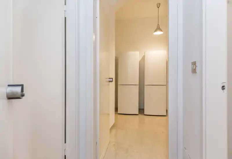 View through a doorway into a small kitchen/storage area with two upright refrigerators under a pendant light; minimal styling and neutral finishes.