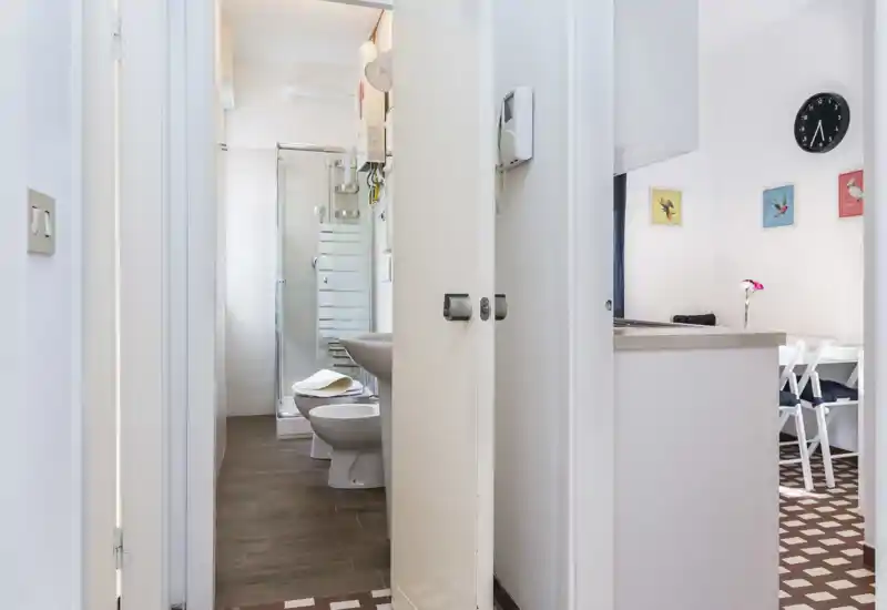 Bright, compact bathroom seen through an open doorway, featuring a shower cabin, sink and bidet; framed with the hallway and a glimpse of the dining nook.
