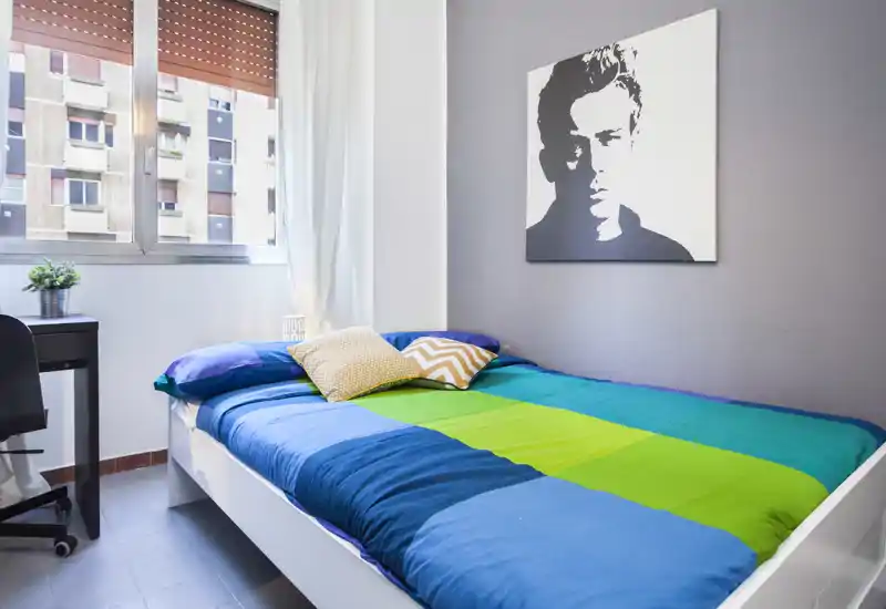 Bright, modern bedroom with a colorful double bed, decorative cushions and a desk by the window. Large window provides natural light and a simple, contemporary wall artwork adds character.