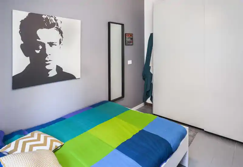 Bright, modern bedroom with a colorful double bed, decorative cushions and a large black-and-white portrait on the wall. Clean lines, a full-height wardrobe and a slim mirror complete the functional layout.