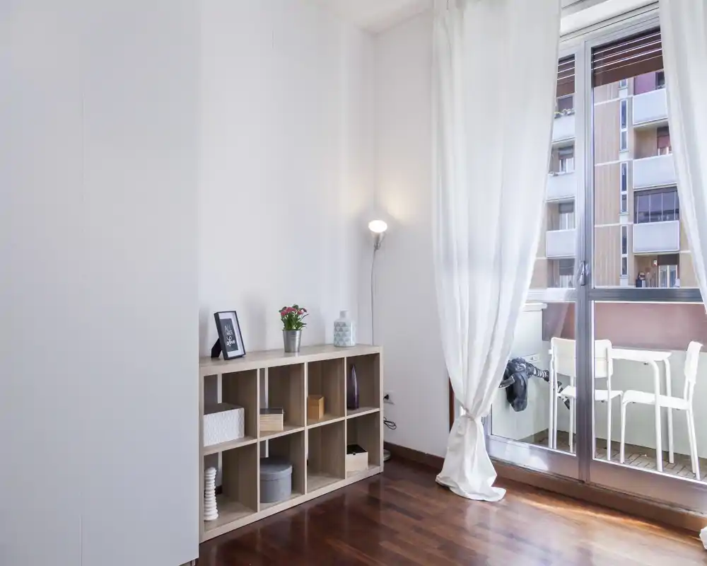Zona giorno luminosa e minimale con pavimento in legno, mobile basso a cubi, lampada da terra e grandi porte scorrevoli che si affacciano su un piccolo balcone.