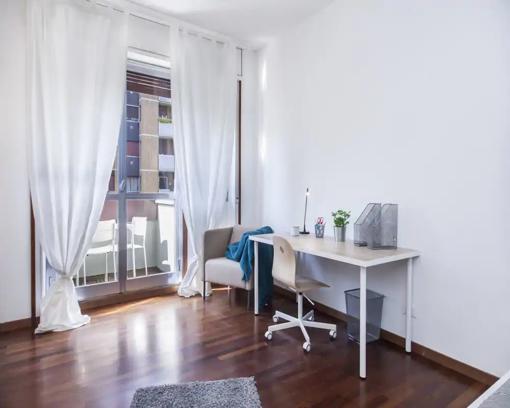 Spazio living luminoso e minimal con una scrivania vicino alla porta finestra verso il balcone, una piccola poltrona e pavimento in legno caldo — ideale come angolo studio con luce naturale.