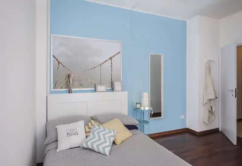 Camera da letto luminosa e di stile con una testiera bianca su una parete azzurra, cuscini decorativi e un comodino con lampada — uno spazio notte accogliente e ben allestito.