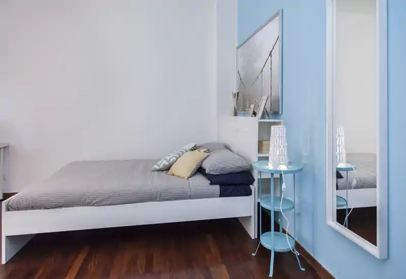 Camera da letto moderna e pulita con struttura letto bianca, biancheria a righe e cuscini decorativi; parete azzurra e specchio a tutta altezza che donano profondità.