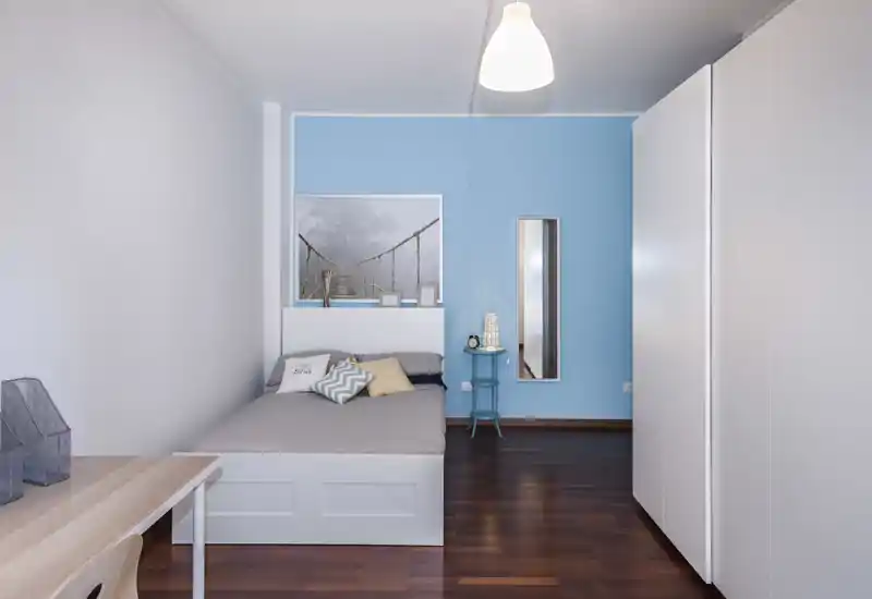 Camera da letto luminosa e moderna con letto matrimoniale contro una parete blu, cuscini decorativi, comodino e ampia armadio; linee pulite e pavimento in legno scuro creano un ambiente calmo e confortevole.