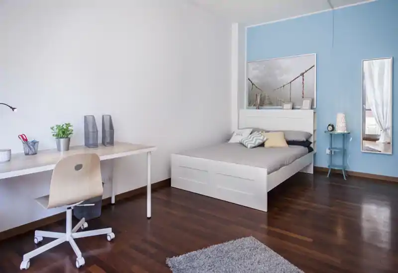 Camera da letto moderna e luminosa con letto bianco, biancheria morbida e cuscini decorativi; sulla sinistra una postazione di lavoro pulita con scrivania e sedia, con quadri e uno specchio che rendono l'ambiente elegante.
