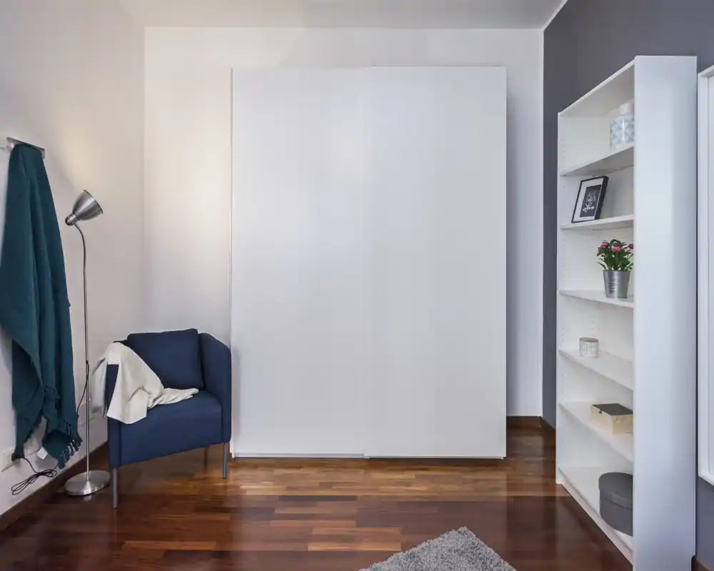 Bright, minimal bedroom corner with a large white wardrobe, shelving unit and a cozy navy armchair — clean lines and warm wooden flooring create a calm atmosphere.