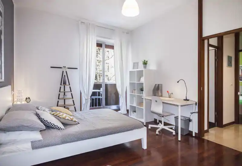 Bright, modern bedroom with a double bed, decorative cushions and a small work desk by the window leading to a balcony. Clean lines, wooden floor and white walls create a calm, welcoming atmosphere ideal for rentals.