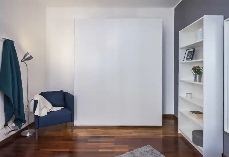 Bright, minimal bedroom corner with a large white wardrobe, shelving unit and a cozy navy armchair — clean lines and warm wooden flooring create a calm atmosphere.