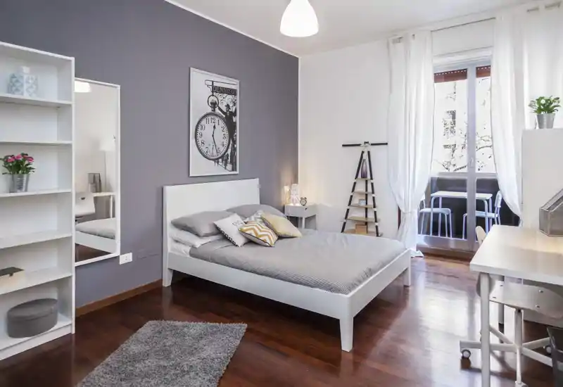 Bright, modern bedroom with a white bed frame, bedside table and desk by a balcony; warm wood floors and tasteful decor create a welcoming atmosphere.