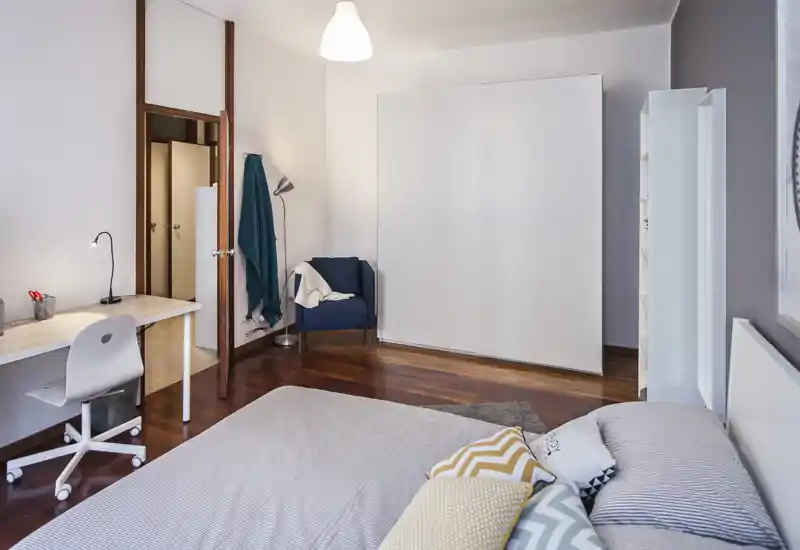 Bright, well-staged bedroom with a neatly made bed in the foreground, a study desk to the left and a large white wardrobe against the wall — calm, modern décor suitable for rentals.