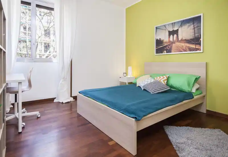 Dormitorio moderno y luminoso con cama doble, cojines coloridos y luz natural procedente de una gran ventana; suelo de madera y cuadro decorativo que aportan calidez.