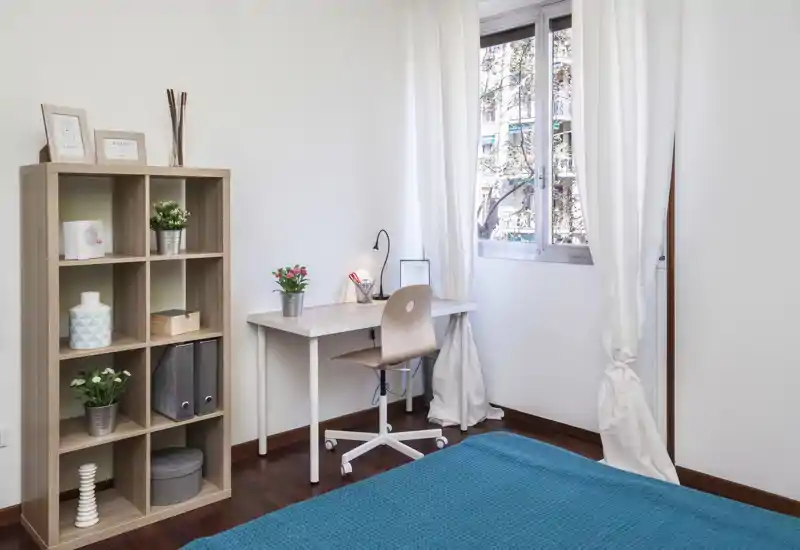 Dormitorio luminoso y ordenado con colcha turquesa, un escritorio junto a la ventana y una estantería de madera: bien iluminado y decorado para descansar y estudiar.