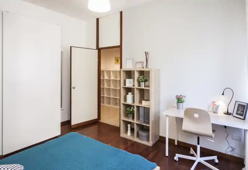 Dormitorio luminoso y ordenado con colcha turquesa, suelo de madera, escritorio pequeño y estantería — ideal para una persona o estudiante.
