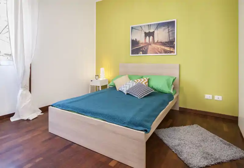 Dormitorio moderno y luminoso con cama de plataforma de madera, cojines coloridos y colcha turquesa frente a una pared de acento verde. La luz natural y el suelo de madera aportan calidez y confort.