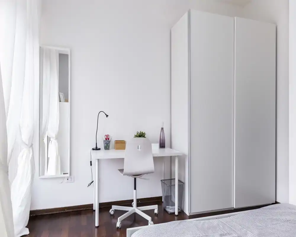 Minimal, bright bedroom corner with a small desk, chair and wardrobe; a slice of the bed is visible in the foreground. Natural light and clean, modern styling create a calm workspace within the sleeping area.