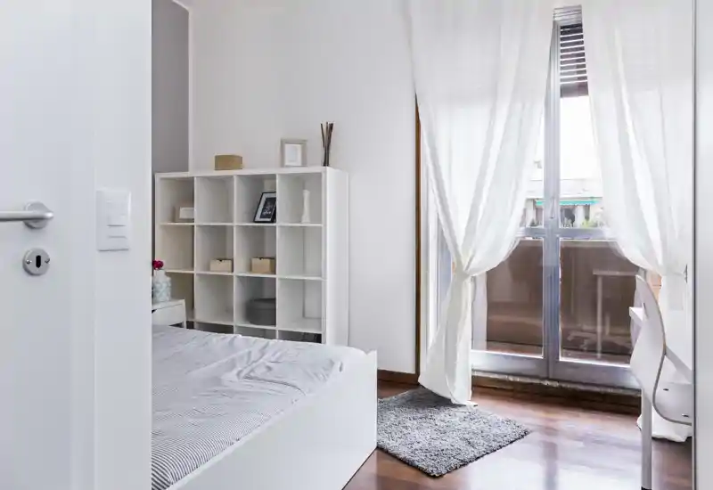 Camera da letto luminosa e moderna con letto bianco, libreria a cubi, morbido tappeto e tende leggere che si aprono su un balcone — stile minimalista e molta luce naturale.