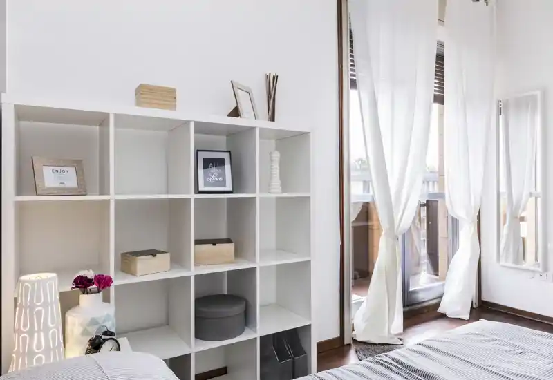 Angolo di camera da letto luminoso e ben arredato con un mobile a cubi, tessili del letto visibili e ampia finestra con tende leggere che lasciano entrare la luce naturale.