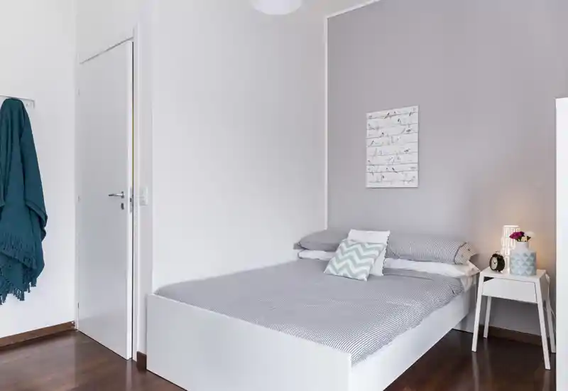 Camera da letto luminosa e minimal con letto matrimoniale ben rifatto, tonalità grigie delicate e un comodino con lampada e decorazioni — perfetta per un soggiorno accogliente.