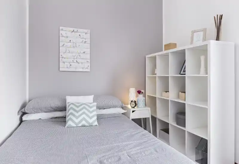 Camera da letto luminosa e minimal con letto matrimoniale ordinato, cuscino decorativo a chevron e libreria a cubi che offre spazio e stile.