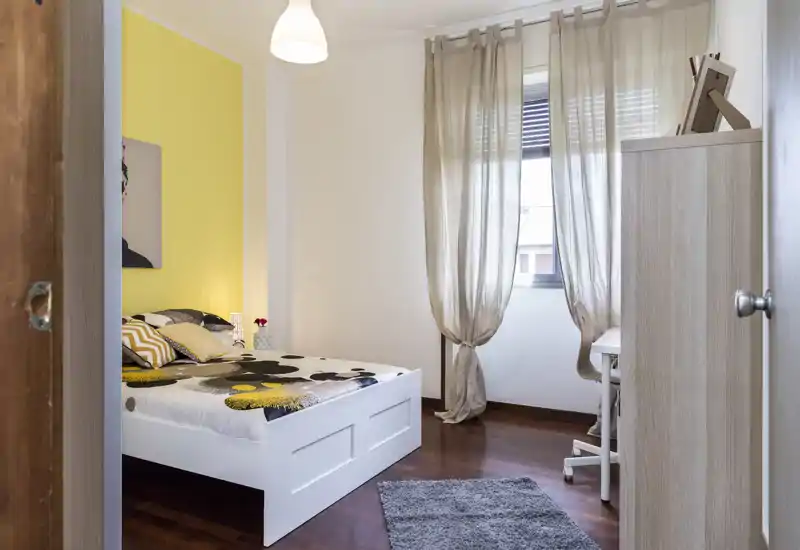 Dormitorio luminoso y ordenado con cama blanca, ropa de cama estampada y pared de acento amarilla; la luz natural que entra por las cortinas aporta una atmósfera acogedora.