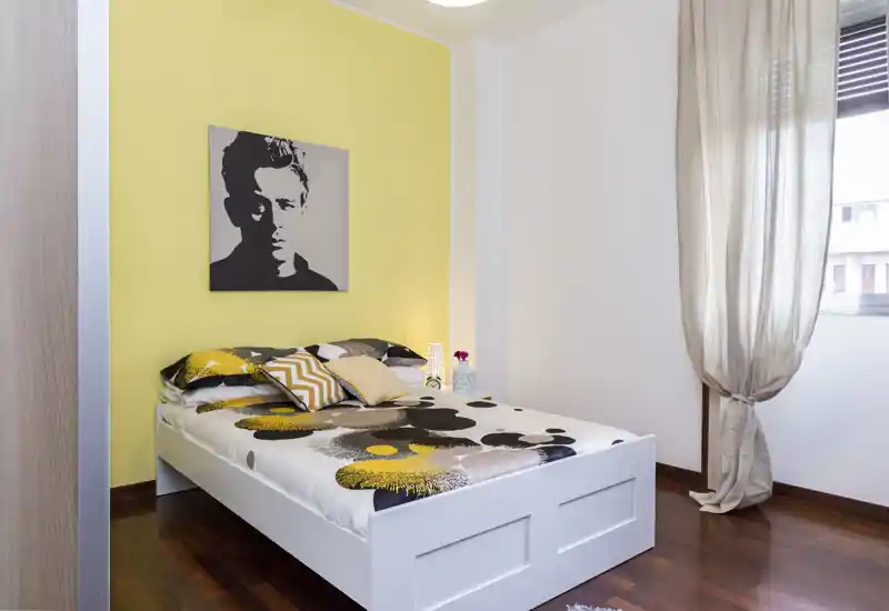 Dormitorio moderno y luminoso con cama blanca, ropa de cama estampada y cojines coordinados frente a una pared amarilla; luz natural y suelos de madera crean un ambiente acogedor.