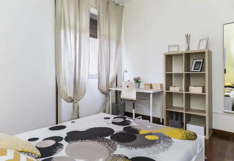 Dormitorio luminoso y bien preparado con colcha estampada, luz natural desde una ventana con cortina y un pequeño escritorio con estantería — ideal para alquileres de corta duración que buscan un espacio para dormir acogedor y funcional.