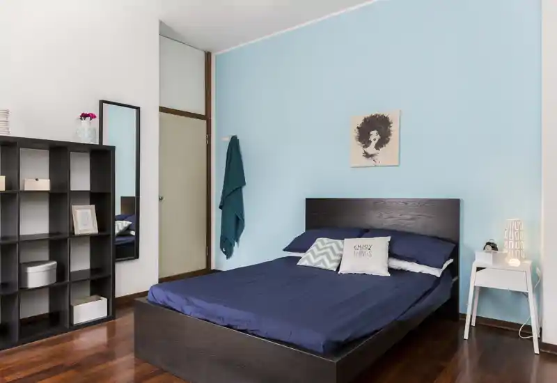 Camera da letto luminosa e curata con letto in legno scuro, biancheria blu navy e cuscini decorativi; parete azzurra e lampada da comodino creano un’atmosfera calma e accogliente.