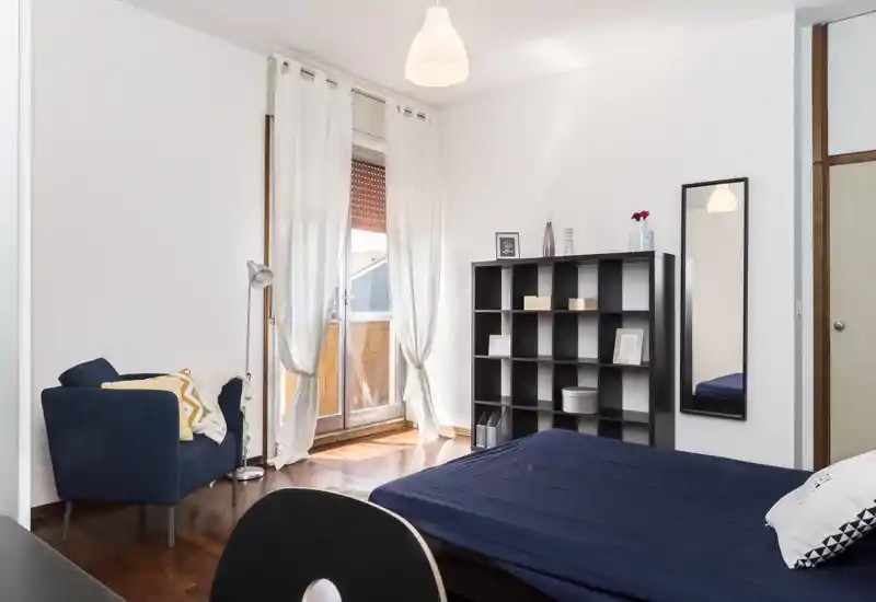 Camera da letto luminosa e moderna con letto matrimoniale e biancheria scura, una poltrona accogliente vicino alla finestra e una libreria a cubi con elementi decorativi. Ampia finestra/porta verso il balcone che illumina bene la stanza.