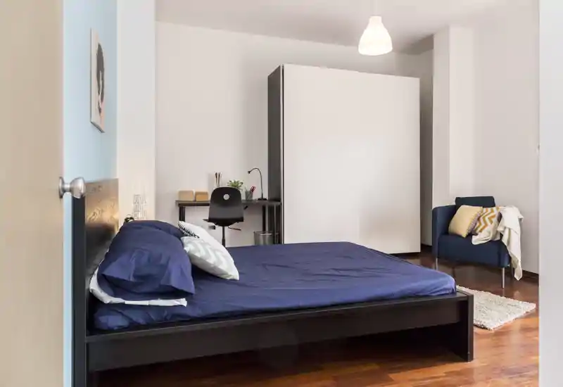 Camera da letto moderna e luminosa con letto basso matrimoniale con biancheria blu, scrivania compatta, armadio e poltrona accogliente — perfetta per un affitto in città.