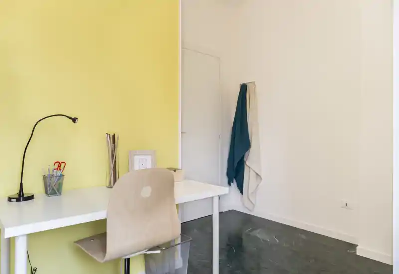 Rincón de estudio ordenado en el salón con escritorio blanco, silla de madera y una lámpara de trabajo frente a una pared amarilla suave; sencillo, luminoso y funcional.