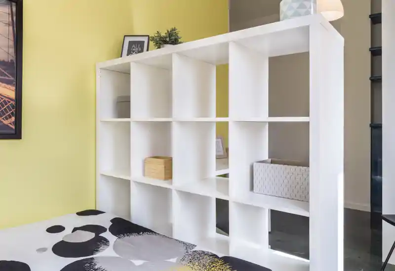Rincón de dormitorio moderno y luminoso con edredón estampado y una estantería blanca modular que separa la zona de descanso del resto del espacio.