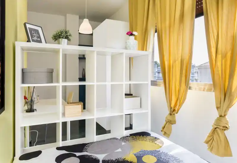 Rincón de dormitorio luminoso y arreglado con cama estampada, estantería blanca de cubos como separador y cortinas amarillas que dejan entrar luz natural.