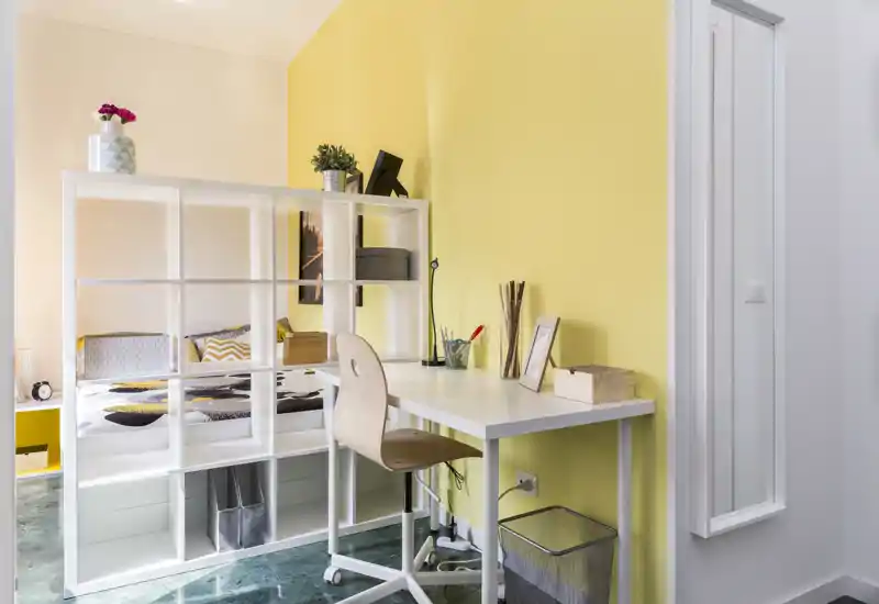 Habitación luminosa y moderna con un separador de estantería blanco y una zona de estudio. La cama se ve detrás de la estantería abierta, con cojines decorativos y detalles amarillos que crean un aspecto fresco y acogedor.