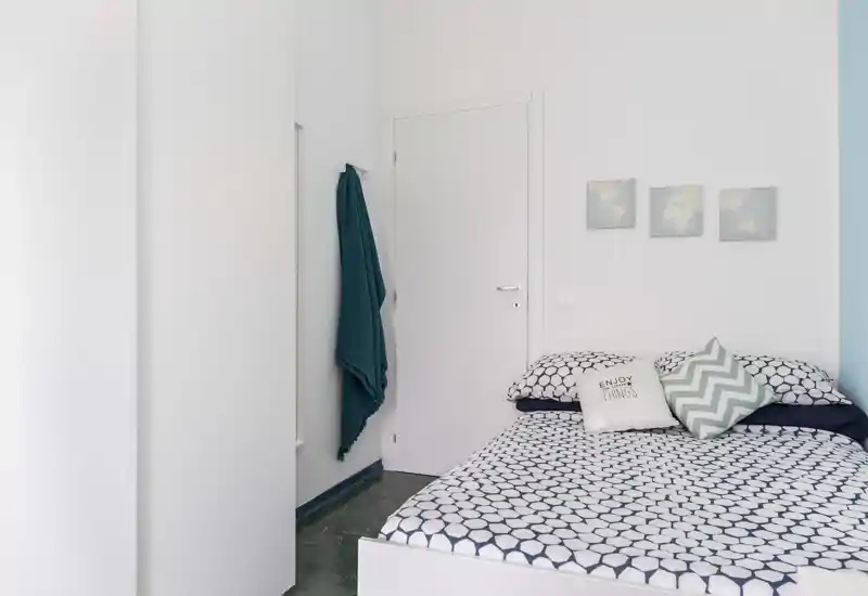 Camera da letto luminosa e minimal con letto matrimoniale a motivo, cuscini decorativi e semplici quadretti alle pareti — ideale per soggiorni accoglienti.