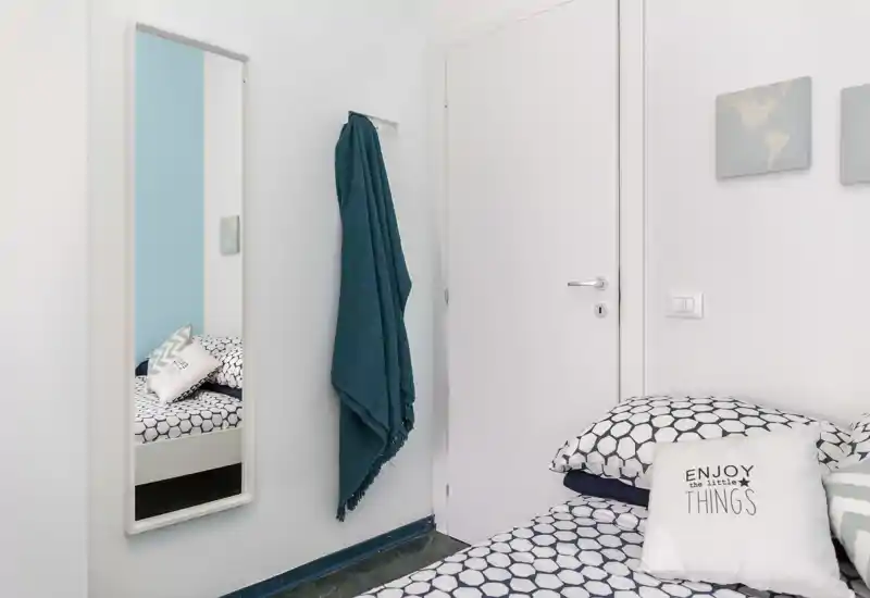 Angolo camera luminoso e ordinato con biancheria a motivo, cuscini decorativi e uno specchio a parete che crea profondità; accenti blu-verdi donano un’atmosfera fresca e rilassante.