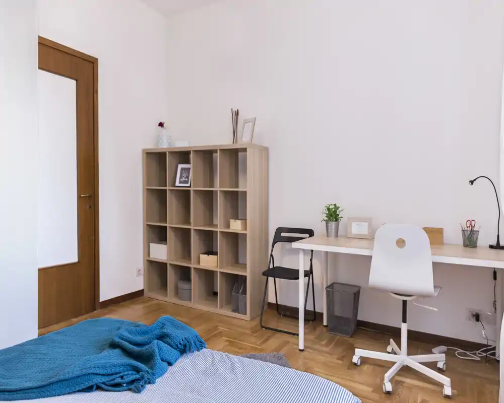 Bright, minimal bedroom with a visible bed corner, wooden shelving and a workspace; pleasant parquet flooring and a cozy blue throw.