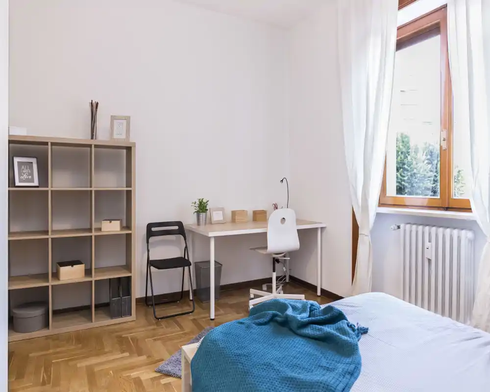 Bright, minimal bedroom with a comfortable bed, desk and shelving — ideal for a student or remote worker. Natural light from the large window and clean, contemporary furnishings create a welcoming space.