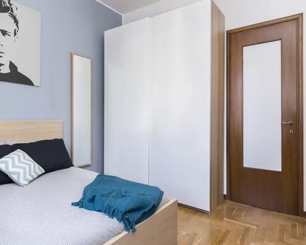 Bright, modern bedroom with a wooden bed, teal throw and a large white wardrobe; calm blue accent wall and mirror add depth.