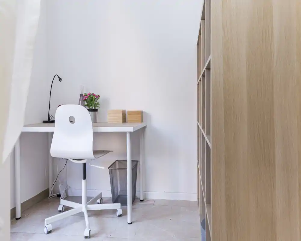 Rincón de trabajo luminoso y minimalista con escritorio blanco y silla con ruedas junto a una estantería; decoración simple con lámpara de escritorio y pequeña planta.