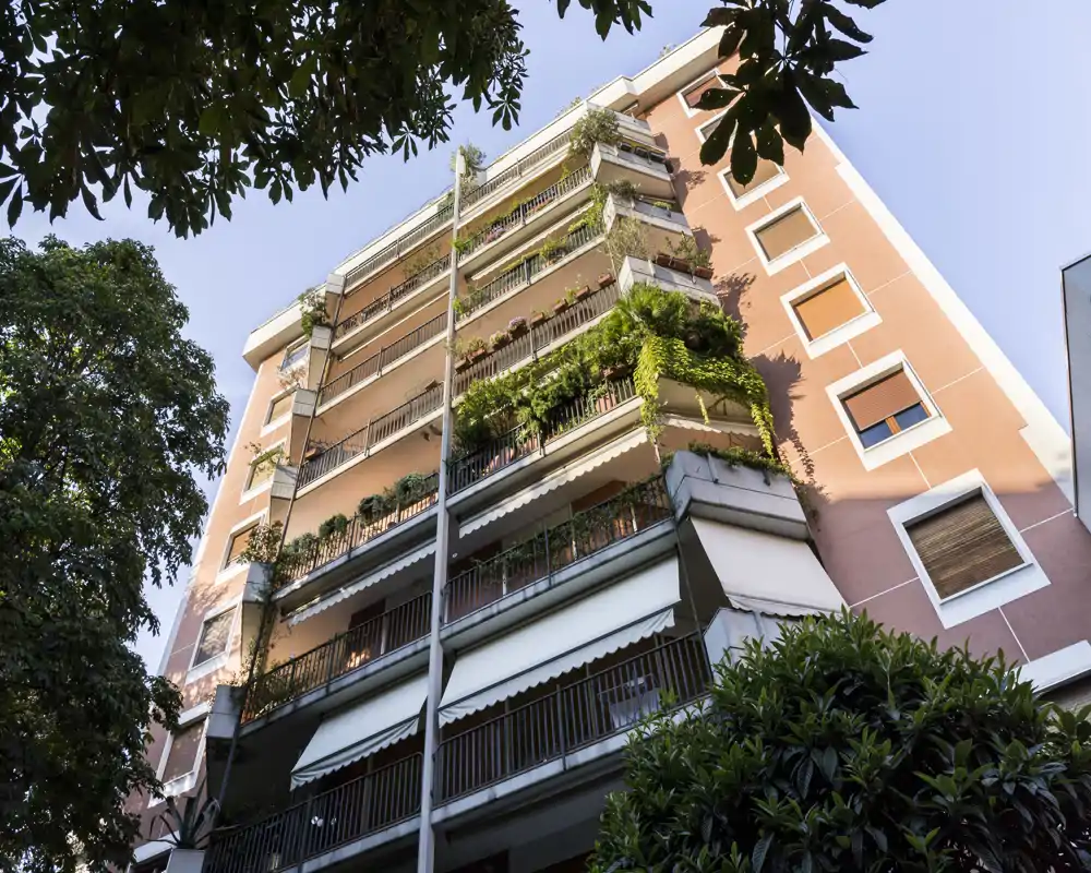 Exterior view of a residential building with multiple balconies and abundant greenery cascading from the terraces, photographed from ground level.