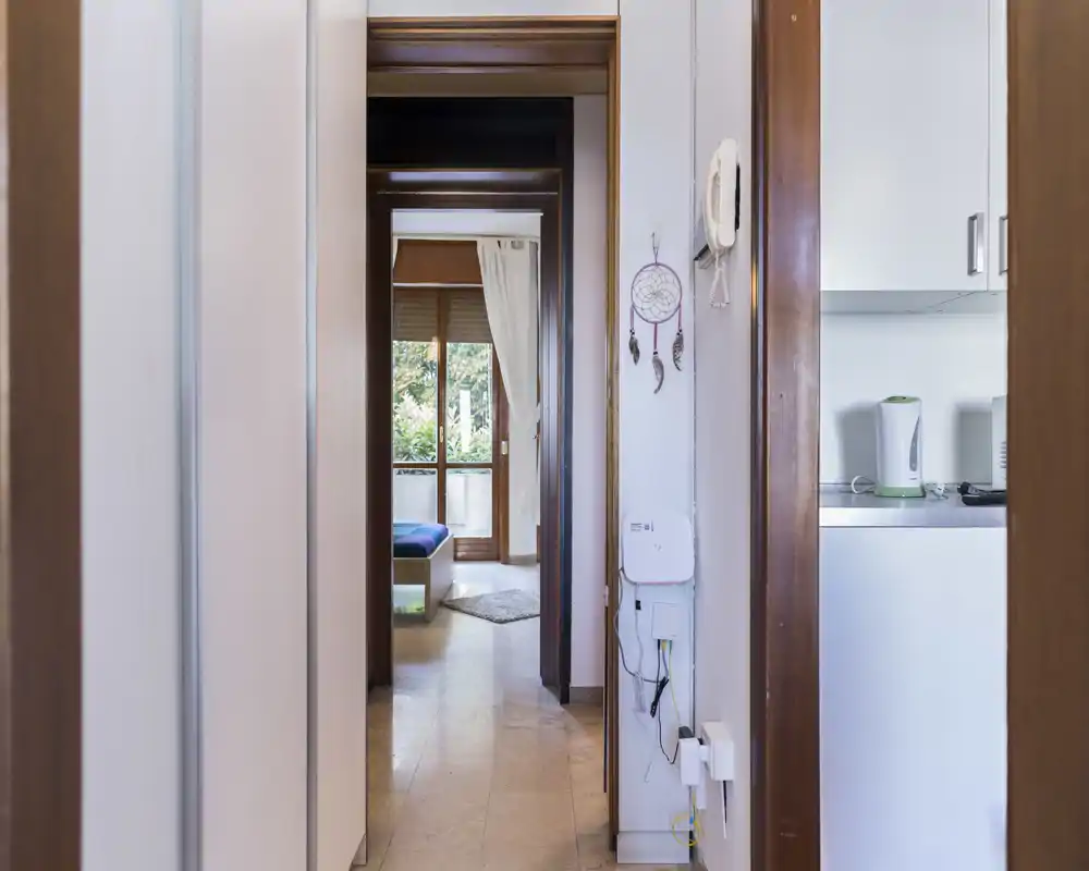 Bright entrance/corridor leading into the apartment with visible doorframes and a glimpse into the living/bed area; clean floors and natural light from the room ahead.
