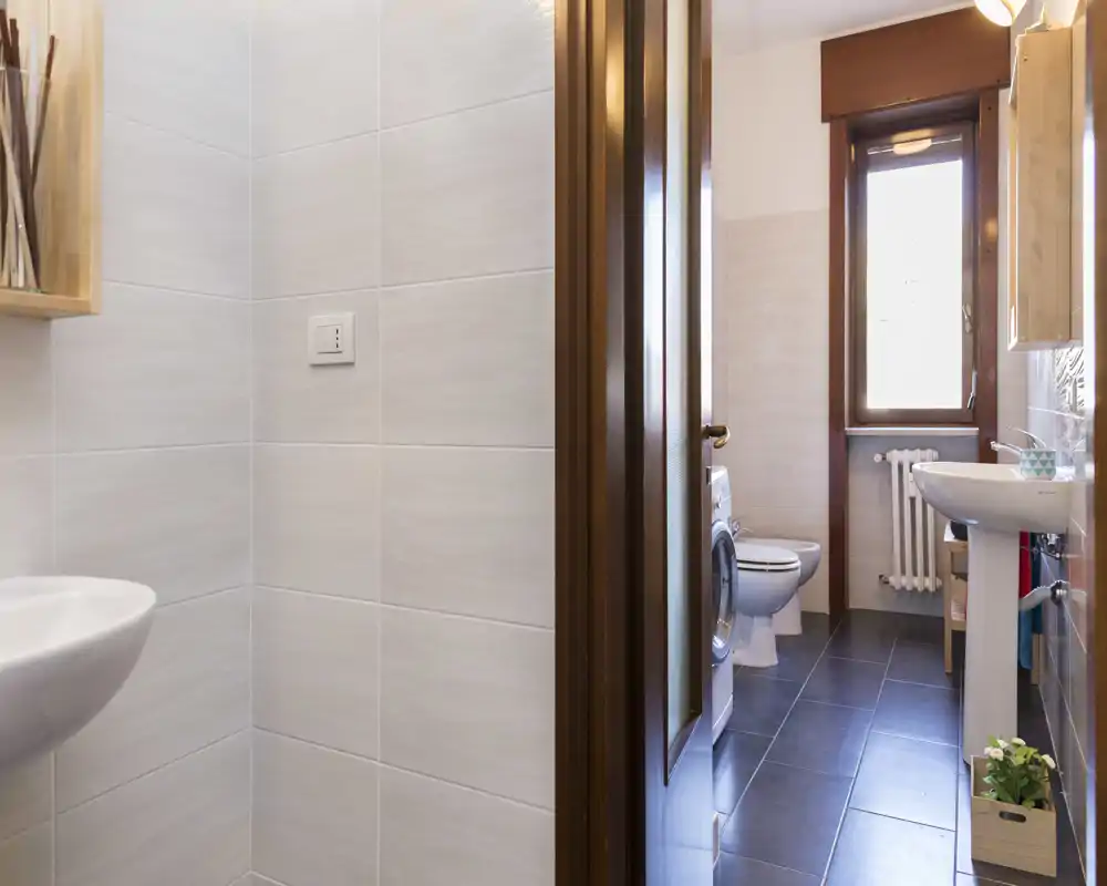Clean, modern bathroom with a sink, toilet and a window providing natural light. The space is tidy with dark floor tiles and white fixtures.