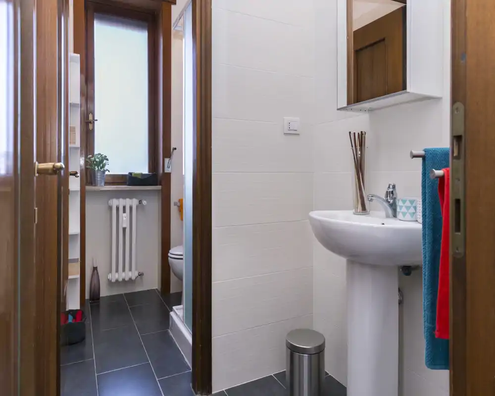 Compact, modern bathroom with a pedestal sink, wall mirror, towel rack and visible toilet in a separate alcove; bright, clean styling suitable for rental listings.