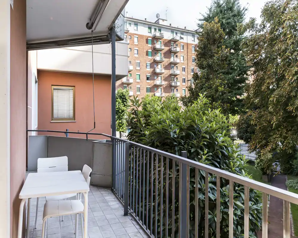 Sunny private balcony with table and chairs, overlooking trees and neighboring buildings — ideal for morning coffee or a small outdoor meal.