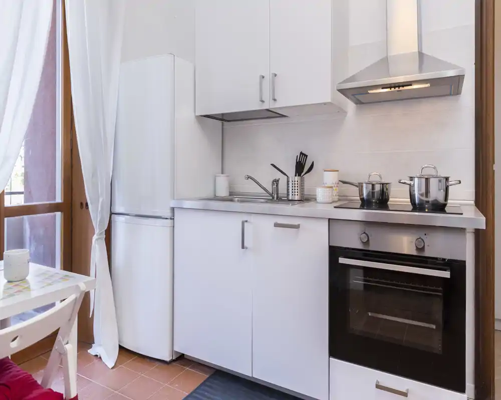 Compact modern kitchen with white cabinets, electric oven and hob, refrigerator and a small dining spot by a bright window — ideal for short stays.